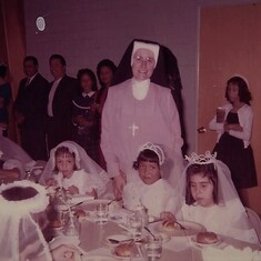 First Communion with her mom, sister and dad standing in the back round by the sisters (nun) shouolder