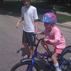 Ethan helping Zoe to ride her bike with one hand and one good leg